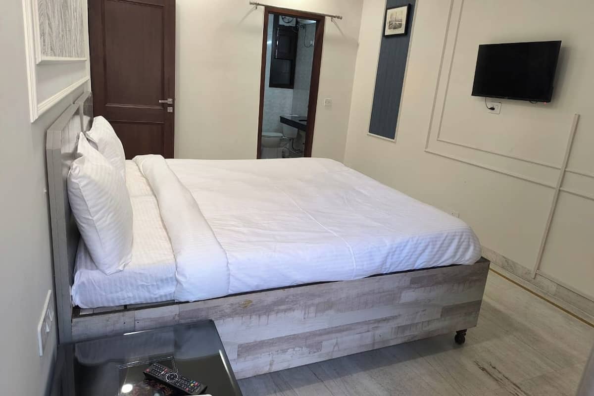 Un dormitorio con cama, mesita de noche con control remoto, cabecera de madera, puerta, espejo, televisor montado en la pared y un pequeño marco de fotos.