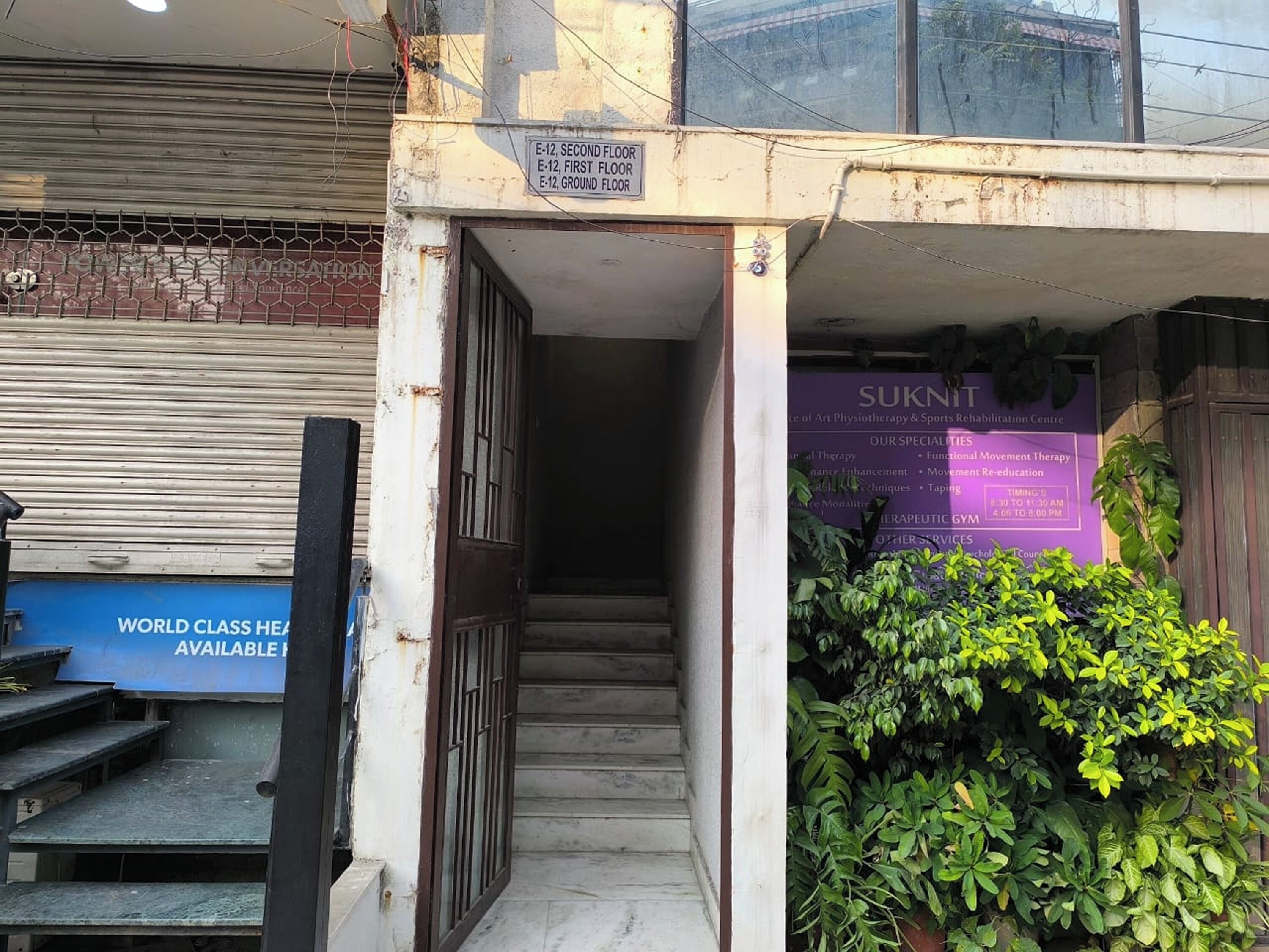 A building entrance with a staircase leading to multiple floors, a closed shutter door, and a sign indicating the ground floor.