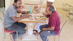 Un coin repas avec une table dressée pour deux personnes, une affiche accrochée au mur et un panneau indiquant l’endroit où se laver les mains.