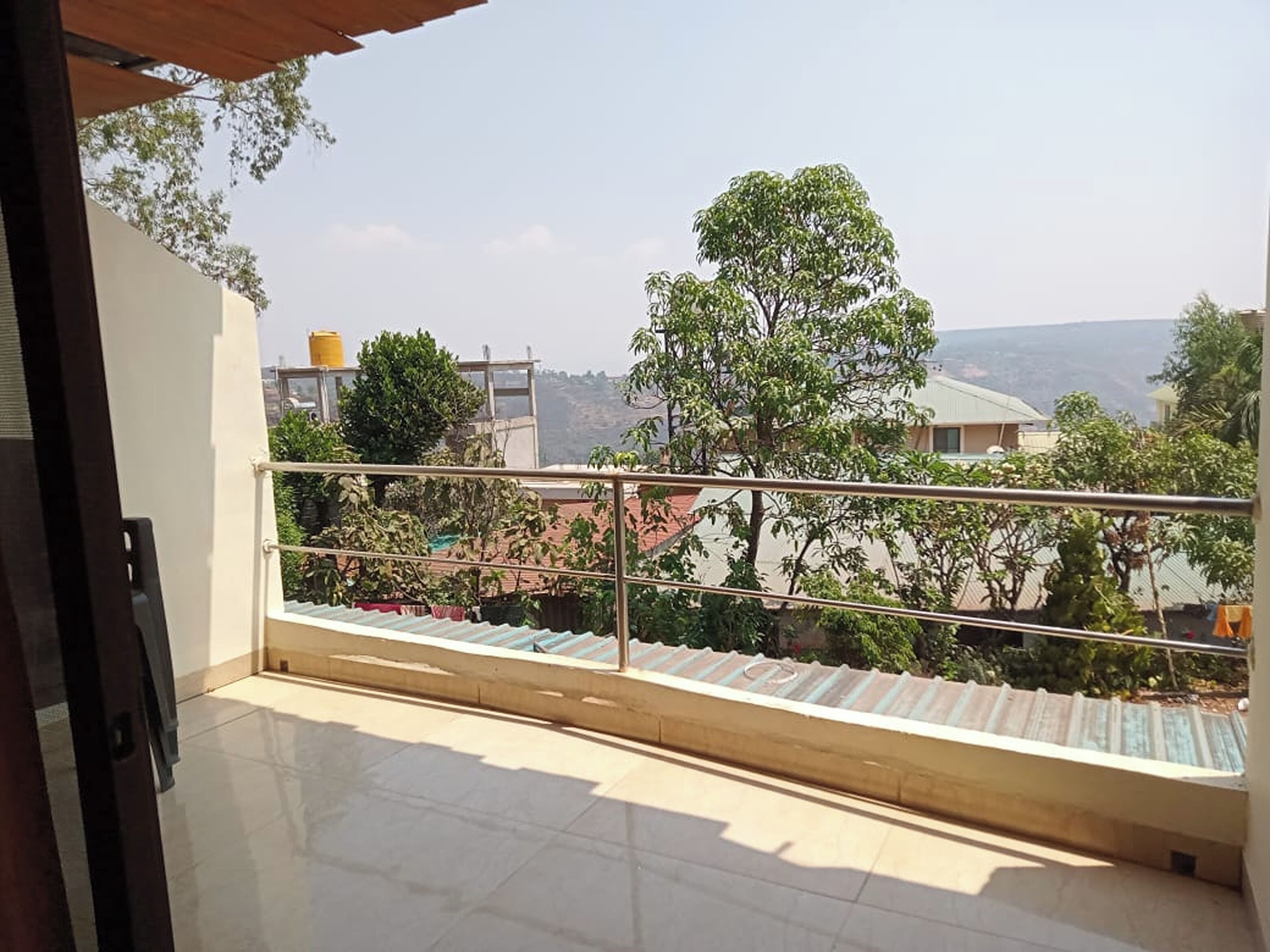 Un balcon doté d’une rambarde en métal, d’un sol carrelé et offrant une vue sur un arbre et des bâtiments au loin.