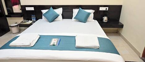 A neatly made bed with white and blue bedding, a bedside table with a kettle, and a trash can.