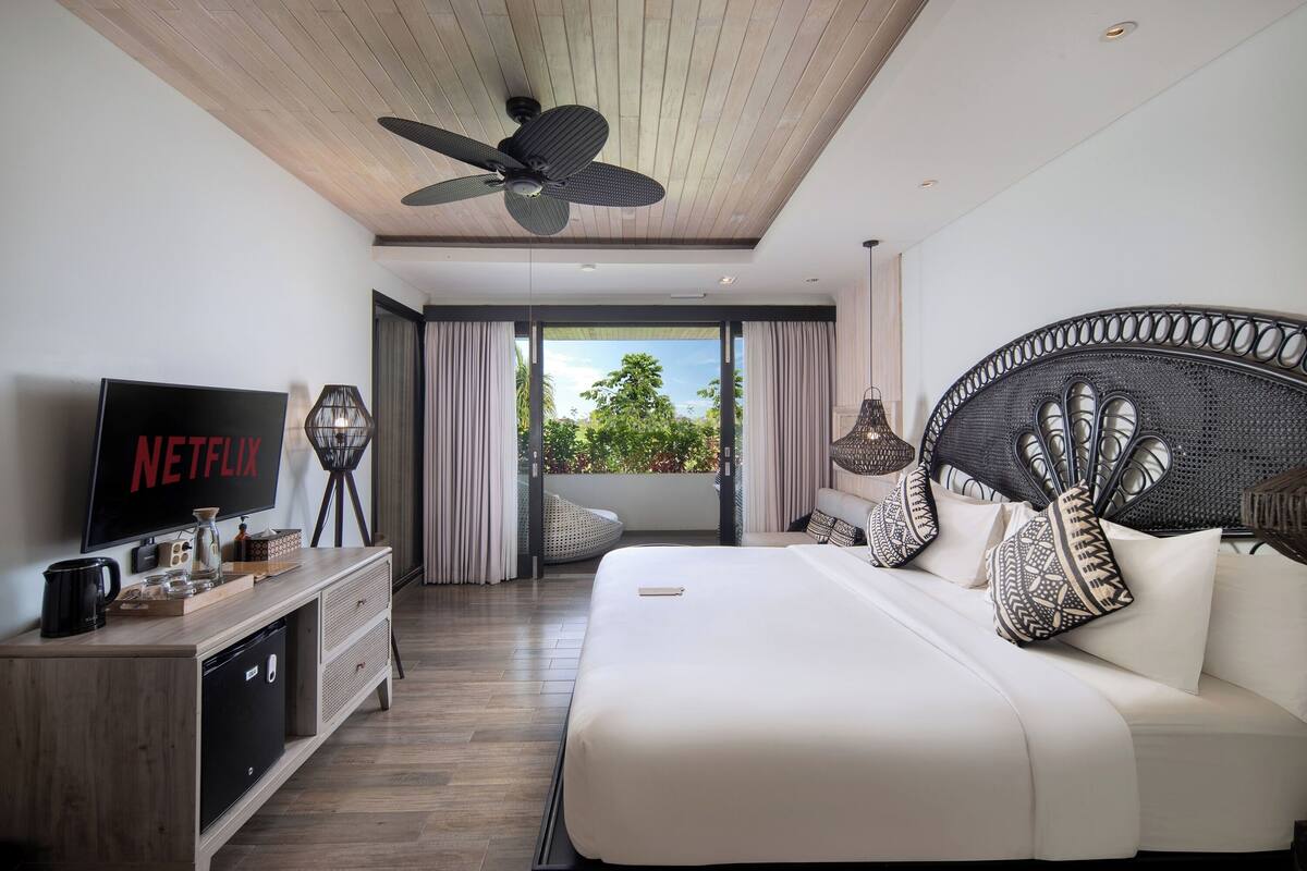 Une chambre d’hôtel moderne avec un grand lit, un téléviseur diffusant du contenu « NETFLIX », un ventilateur de plafond et une vue sur la verdure à travers les portes du balcon.