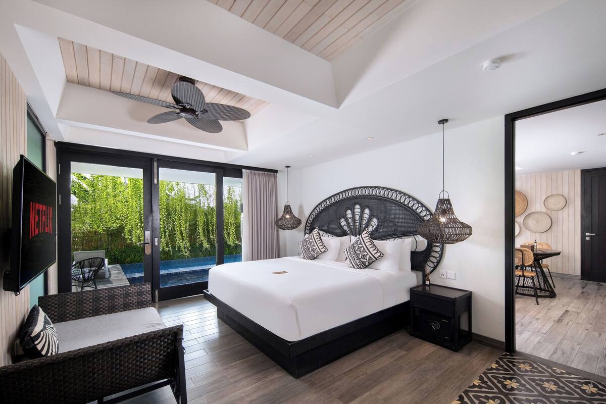 Une chambre moderne avec un grand lit, un ventilateur de plafond, une télévision et un balcon offrant une vue sur la verdure.