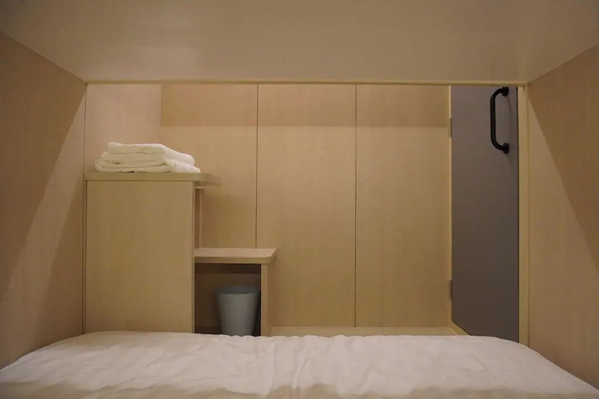 A bedroom with a bed, a wardrobe, a small shelf with towels, and a trash bin.