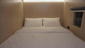 Ein Einzelbett mit weißer Bettwäsche in einem Zimmer mit beige gefärbten Wänden und einem kleinen Fenster mit Jalousie.
