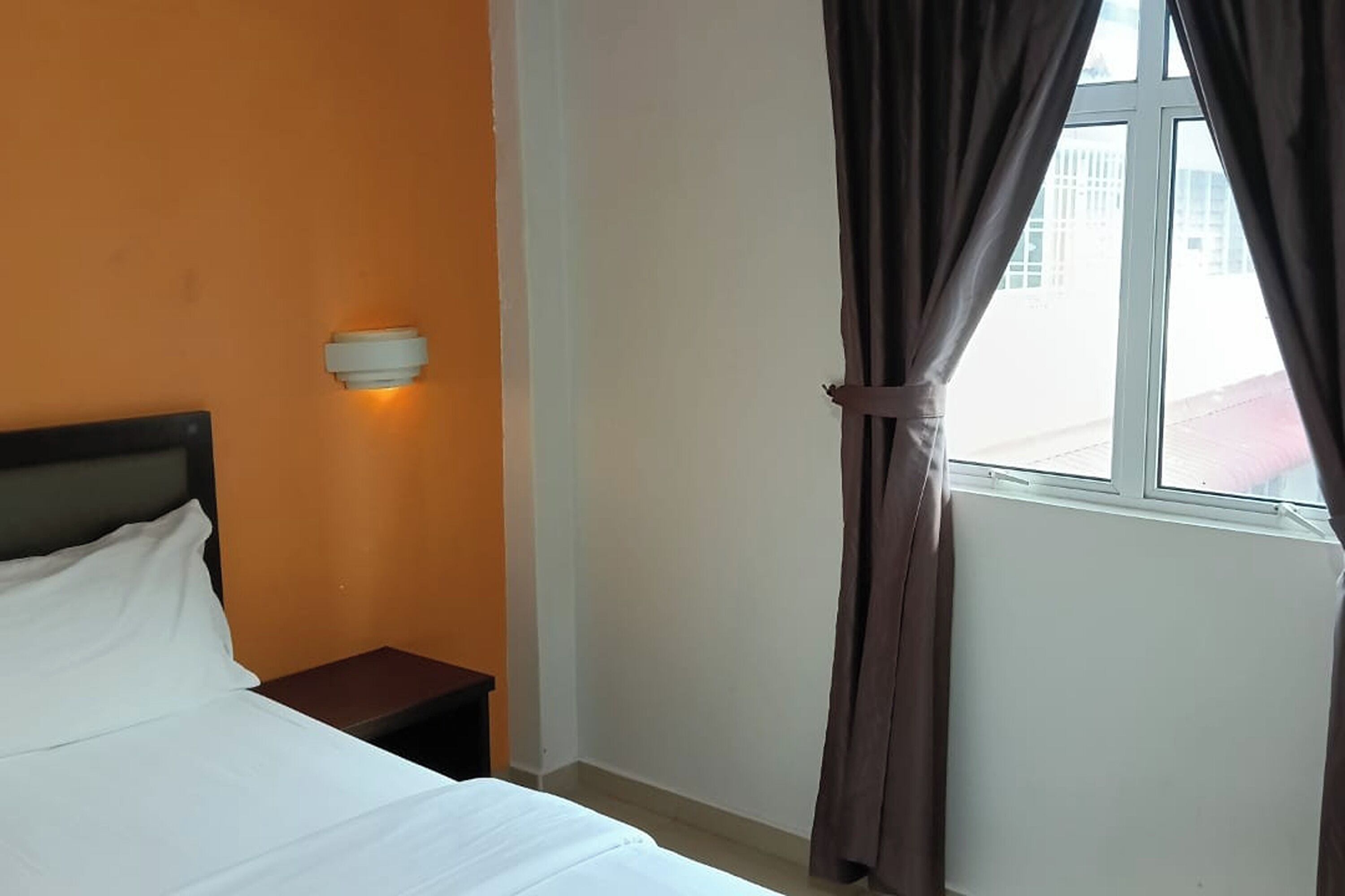 A hotel room with a bed, a nightstand, a wall-mounted light, and a window with curtains.