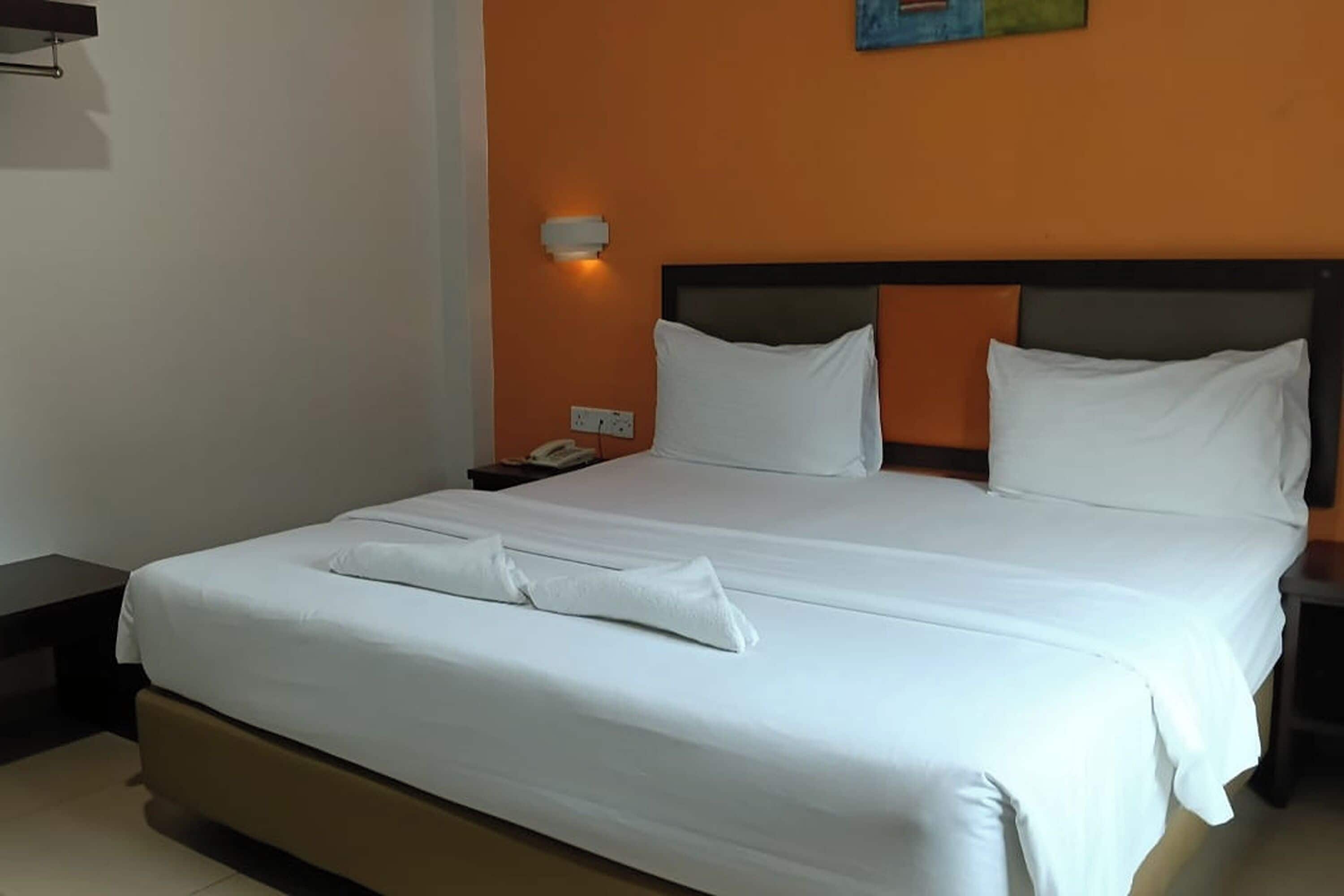 Een net opgemaakt bed met witte lakens en een opgevouwen handdoek, een nachtkastje met een telefoon, en een wandlamp.