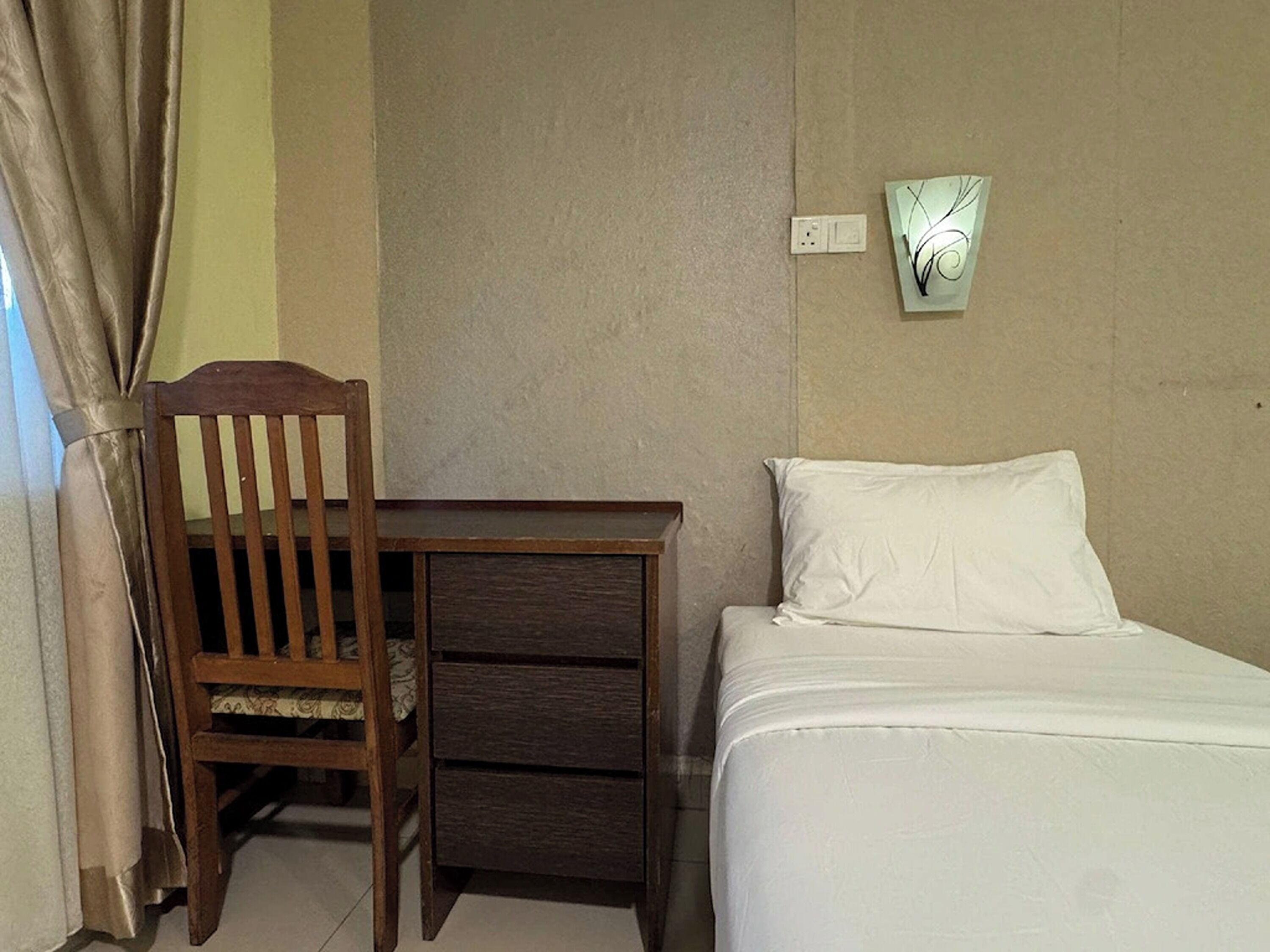 Um quarto com cama, uma cadeira de madeira, uma mesinha e um candeeiro fixado na parede.