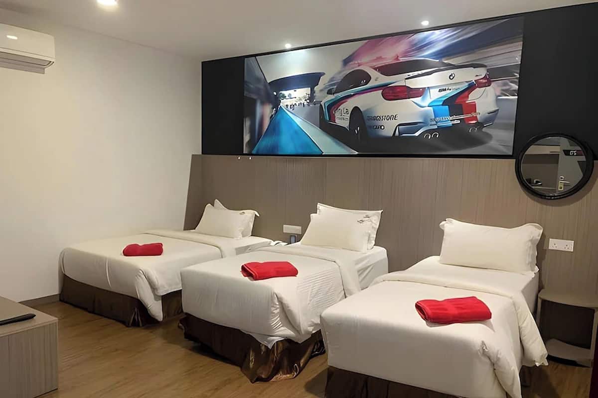 Ein Hotelzimmer mit drei Betten, jedes mit weißer Bettwäsche und einem roten Kissen. Ein flachbildfernseher an der Wand zeigt ein Rennauto.
