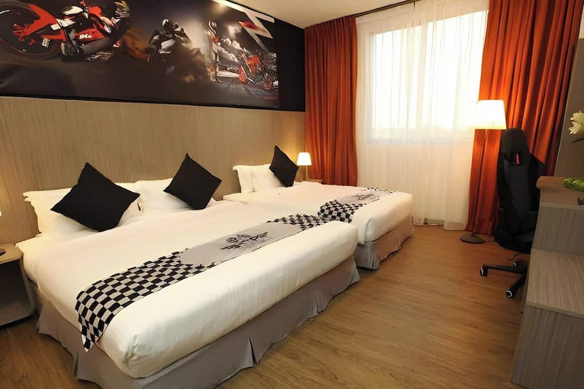 Ein Hotelzimmer mit zwei Betten, einer karierten Tagesdecke, einem großen Wandbild mit Motorradmotiv, einem Schreibtisch mit Lampe und einem Stuhl.