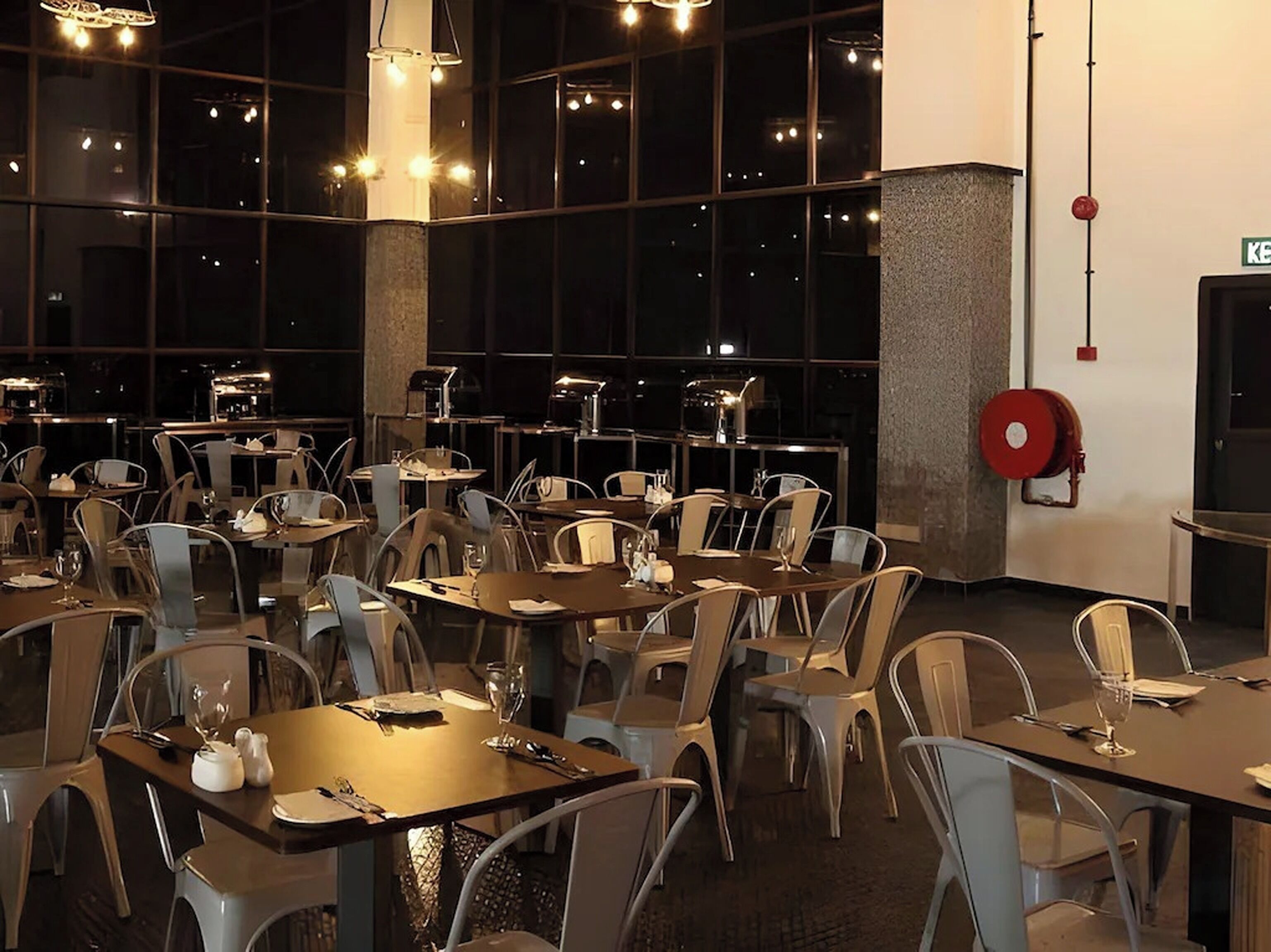 A restaurant with multiple tables and chairs, large windows, and hanging lights.