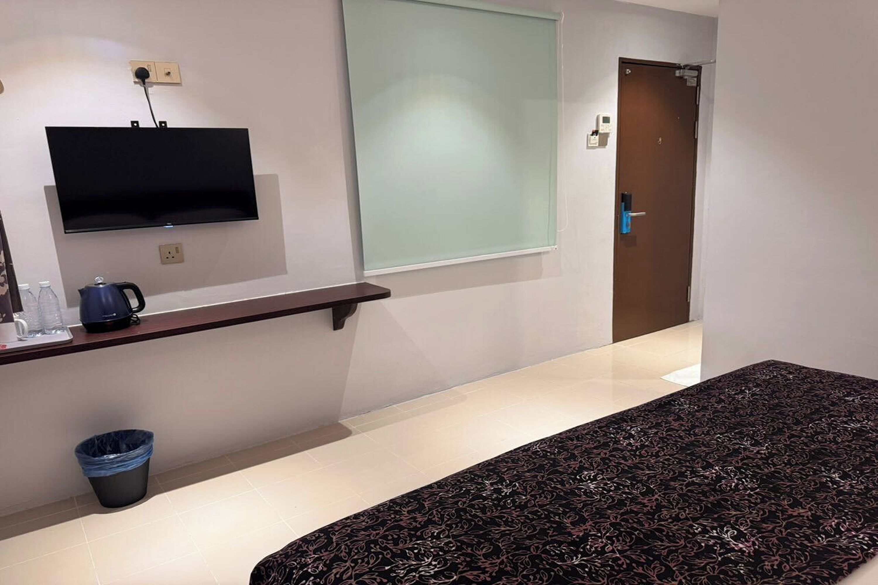 Habitación de hotel con televisor de pantalla plana montado en la pared, una mesita con hervidor y tazas, un cesto de basura y una alfombra estampada.