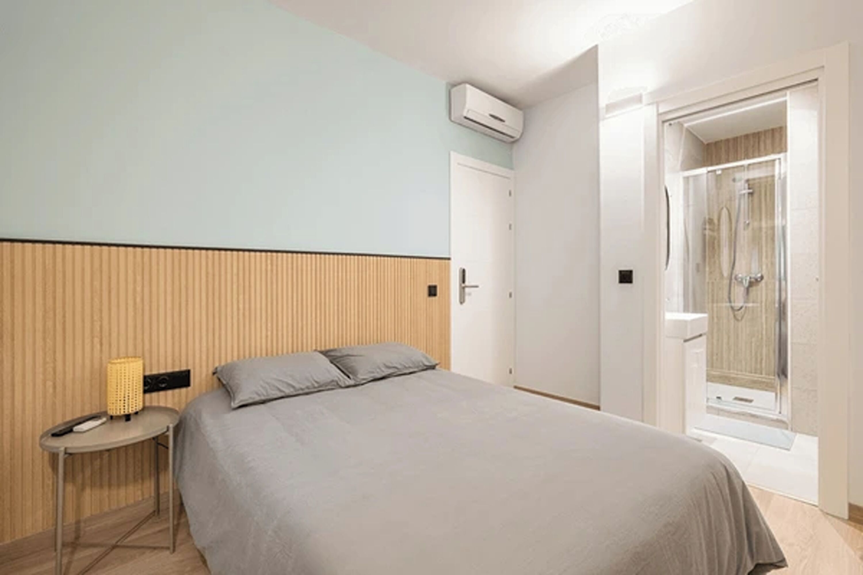 A bedroom with a bed, a bedside table with a lamp, a door, and a shower area.