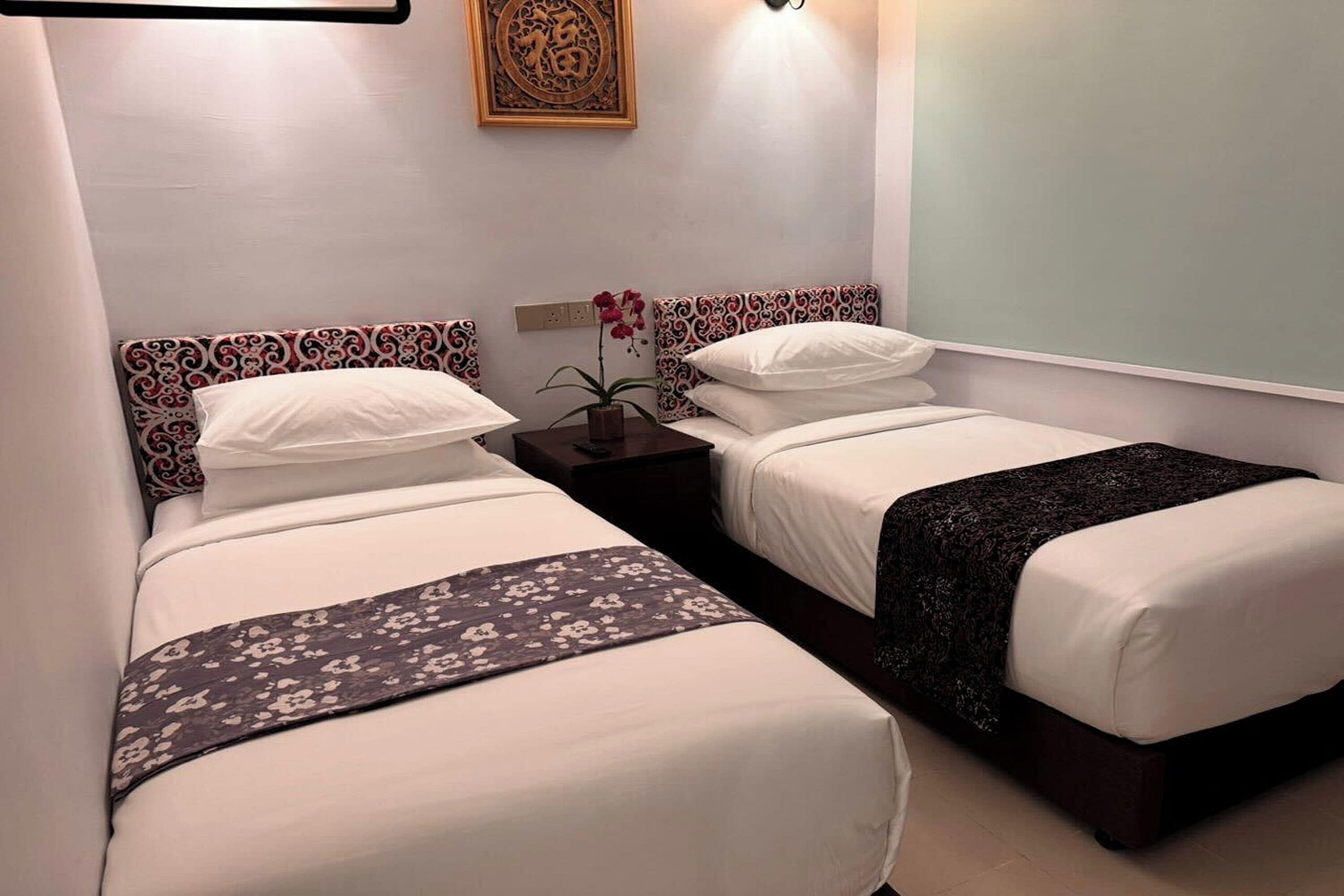 A hotel room with two single beds, a nightstand with a lamp and a vase of flowers, and a decorative wall piece above the beds.