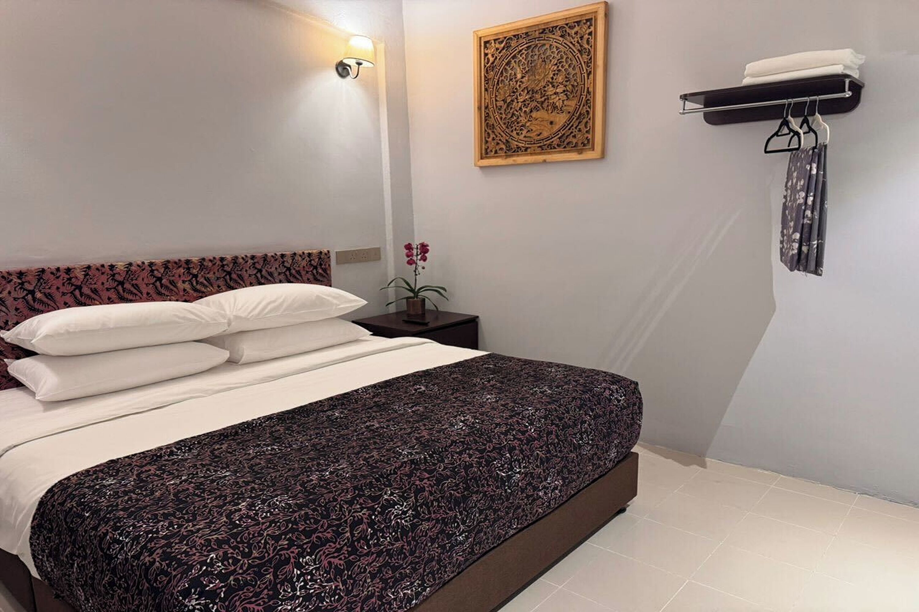 A neatly made bed with a patterned bedspread, a wall-mounted light fixture, a decorative wooden panel, and a towel rack with folded towels.
