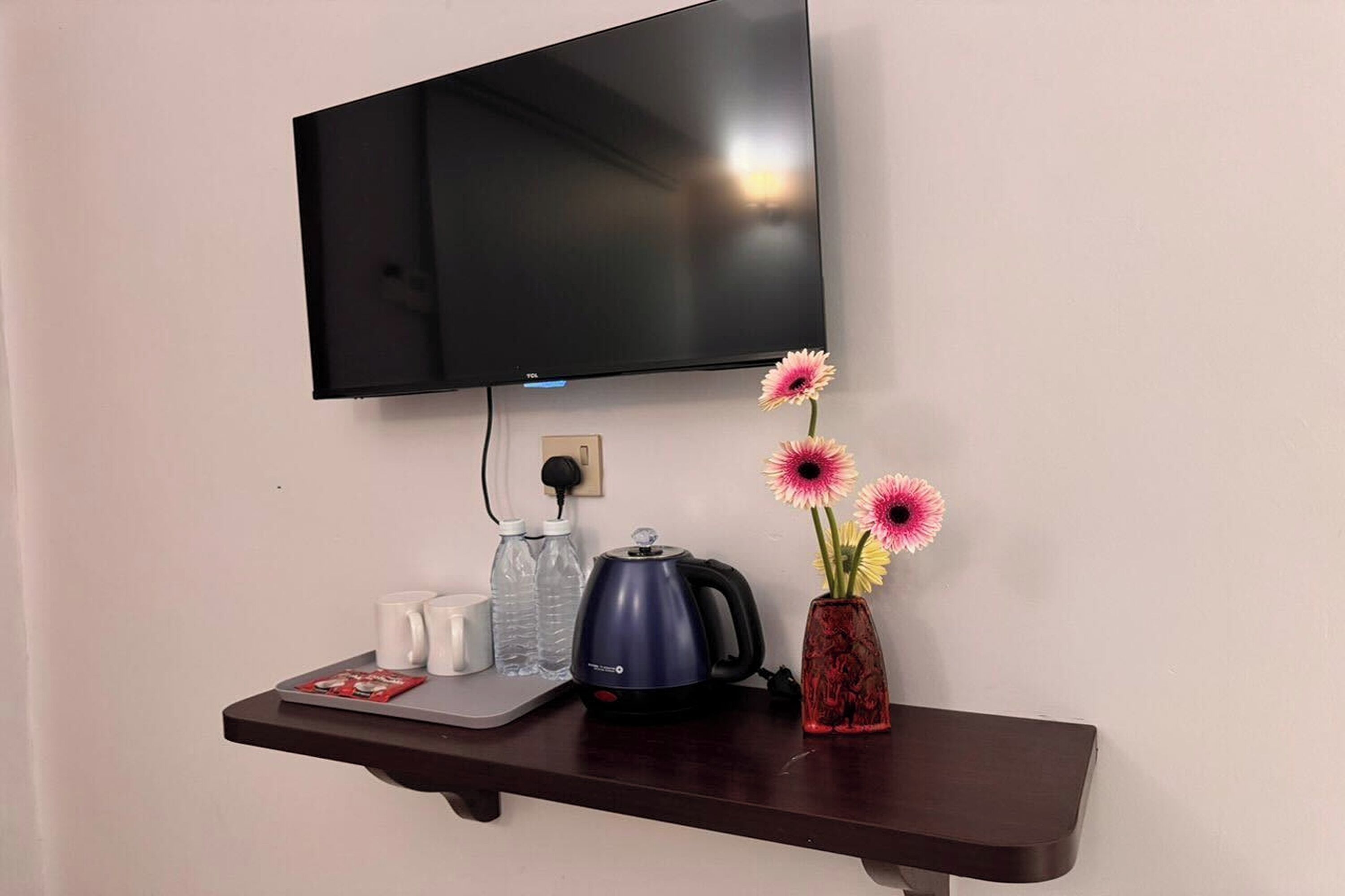 Un televisor montado en la pared sobre una mesita con una tetera, botellas de agua y un florero con flores.