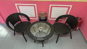 Ein runder Glastisch mit zwei schwarzen Korbstühlen, einem Metallmülleimer und einem roten Feuerlöscher vor einer rosa Wand mit weißen Zierpaneelen.