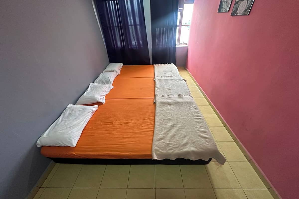 Una cama individual con un colchón naranja y almohadas blancas en una habitación con piso de baldosas y paredes rojas.