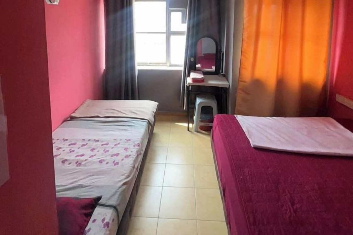 A room with two beds, a window with curtains, a small table with a mirror, and a trash bin.