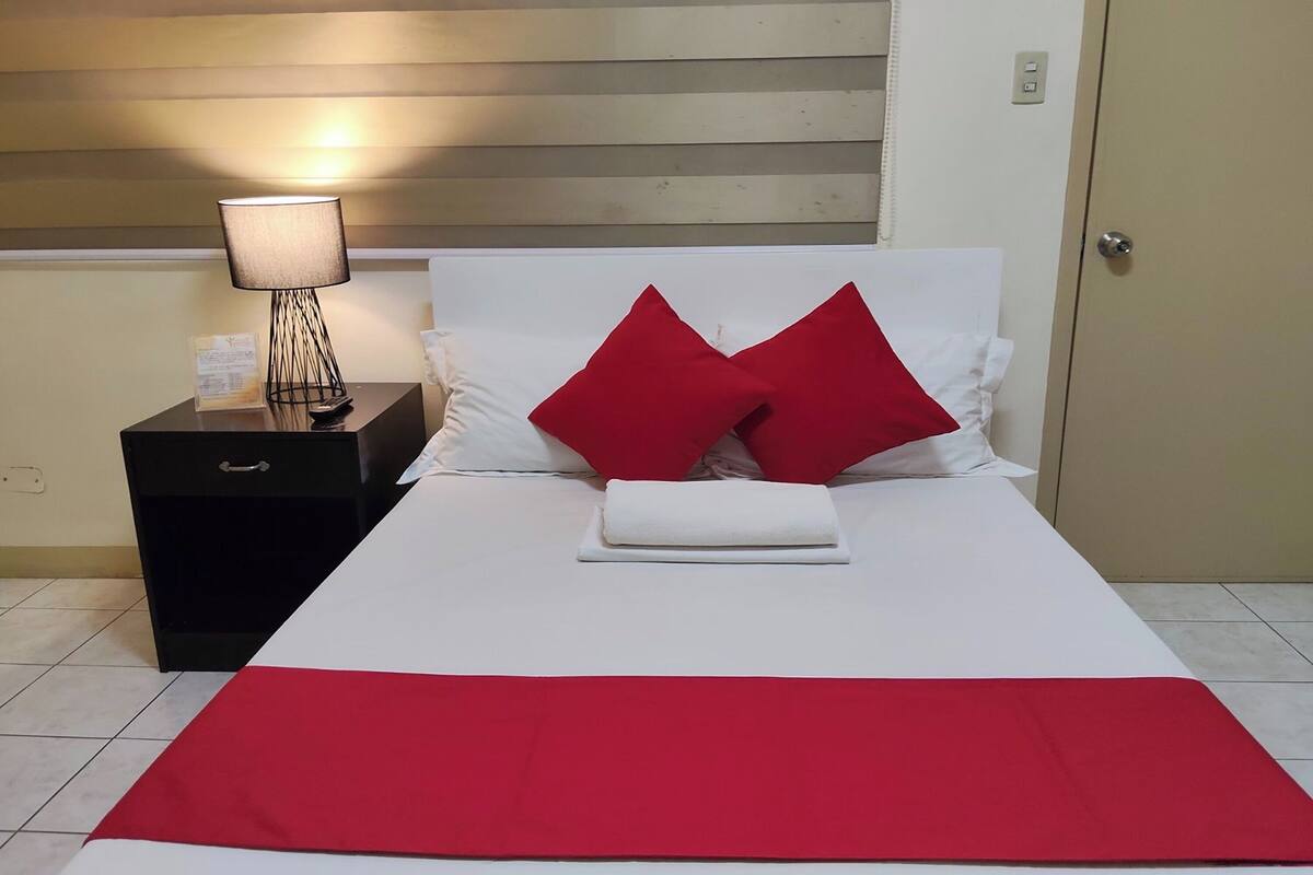 A neatly made bed with white and red bedding, a bedside lamp, and a black nightstand.