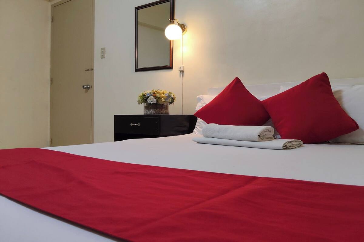 A neatly made bed with red and white bedding, a small bedside table with a vase of flowers, and a wall-mounted mirror.