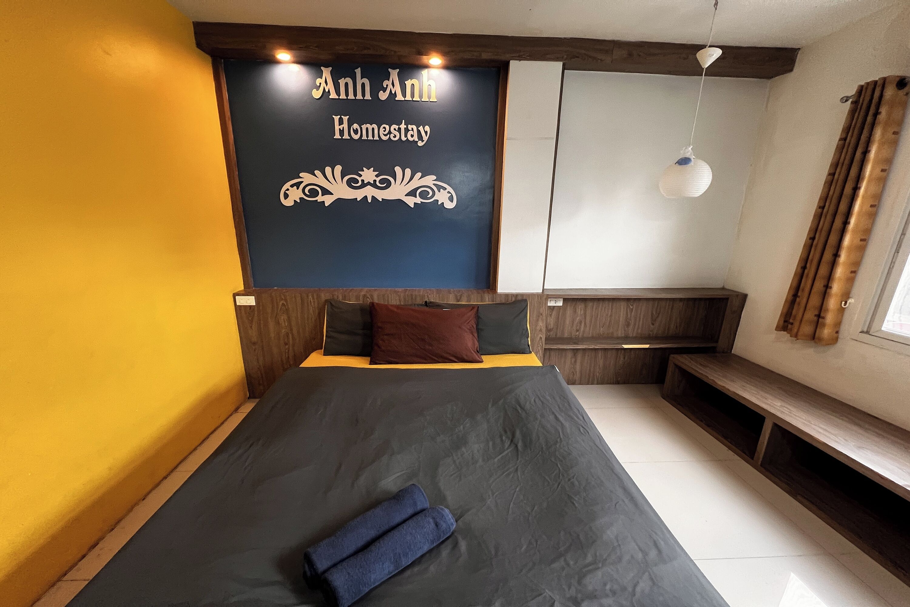 A bedroom with a bed, a bench, and a wall-mounted sign reading "Anh Anh Homestay".