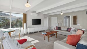 Uma sala de estar moderna com sofá, mesa de centro e televisão. Há uma área de jantar com mesa e cadeiras. O quarto possui janelas amplas e vista para árvores.