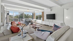 Uma sala de estar espaçosa com um sofá grande, uma mesa de centro e uma TV fixada na parede. Há uma varanda com vista para árvores e um corrimão.