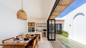 A modern kitchen with a dining area, a built-in oven, and a large window overlooking a pool. - Villa Bellora by Pertama Stay (Seminyak)