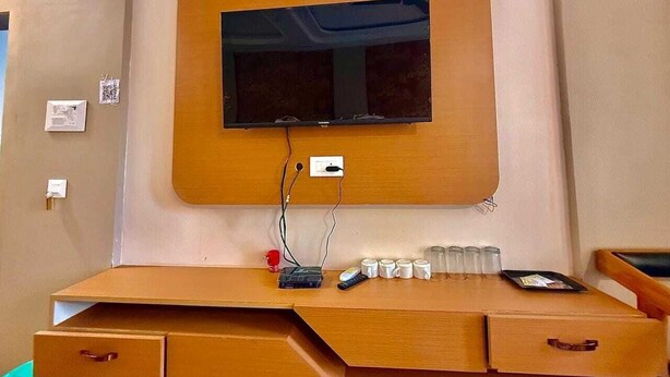 A hotel room with a wooden desk, a flat-screen TV mounted on the wall, a power outlet, and a tray with glasses and a remote control. - The Kridha Palace (Ayodhya)