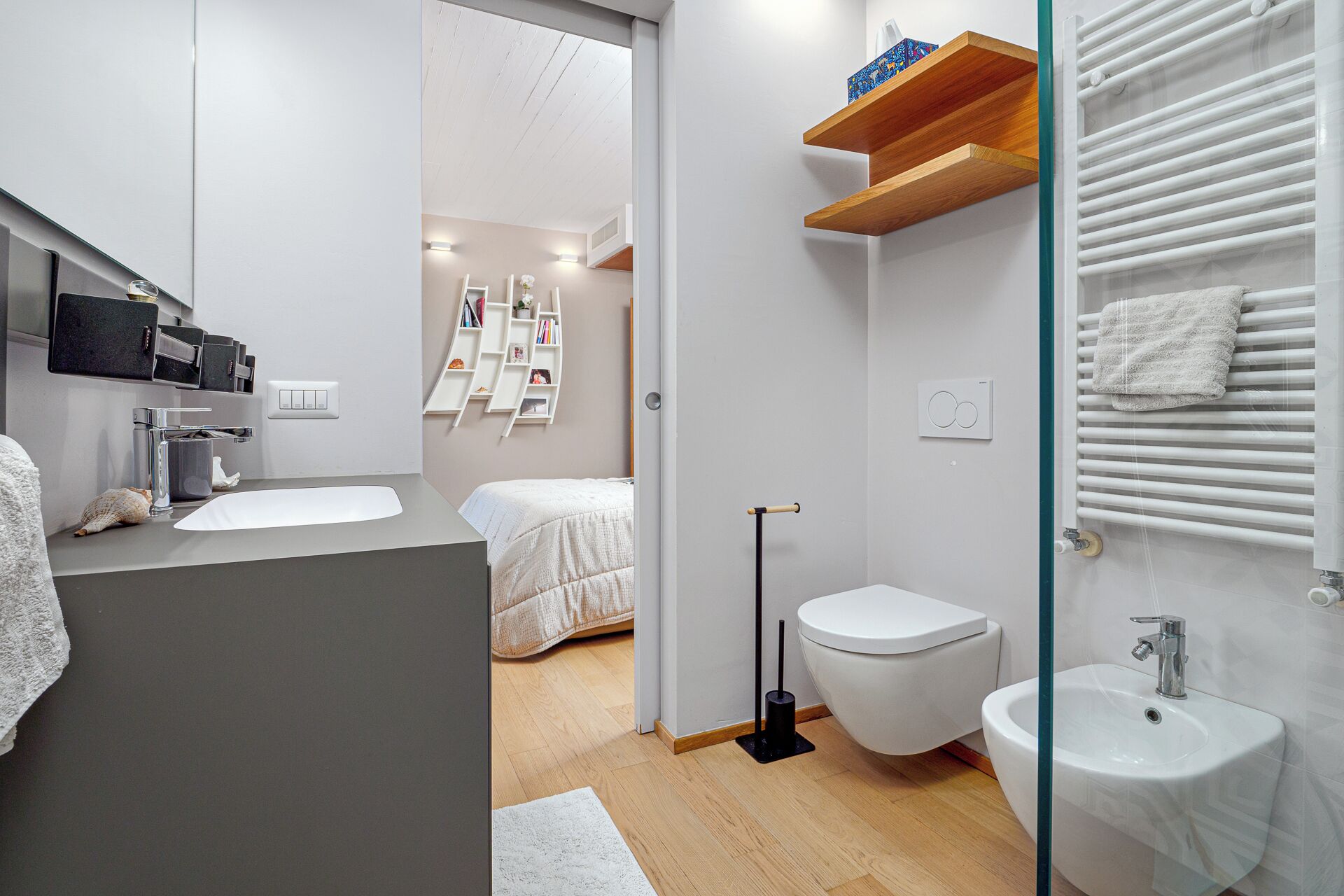 Une salle de bain moderne équipée de toilettes, d’un bidet et d’un lavabo. On trouve une étagère en bois au-dessus des toilettes et un porte-serviettes fixé au mur.