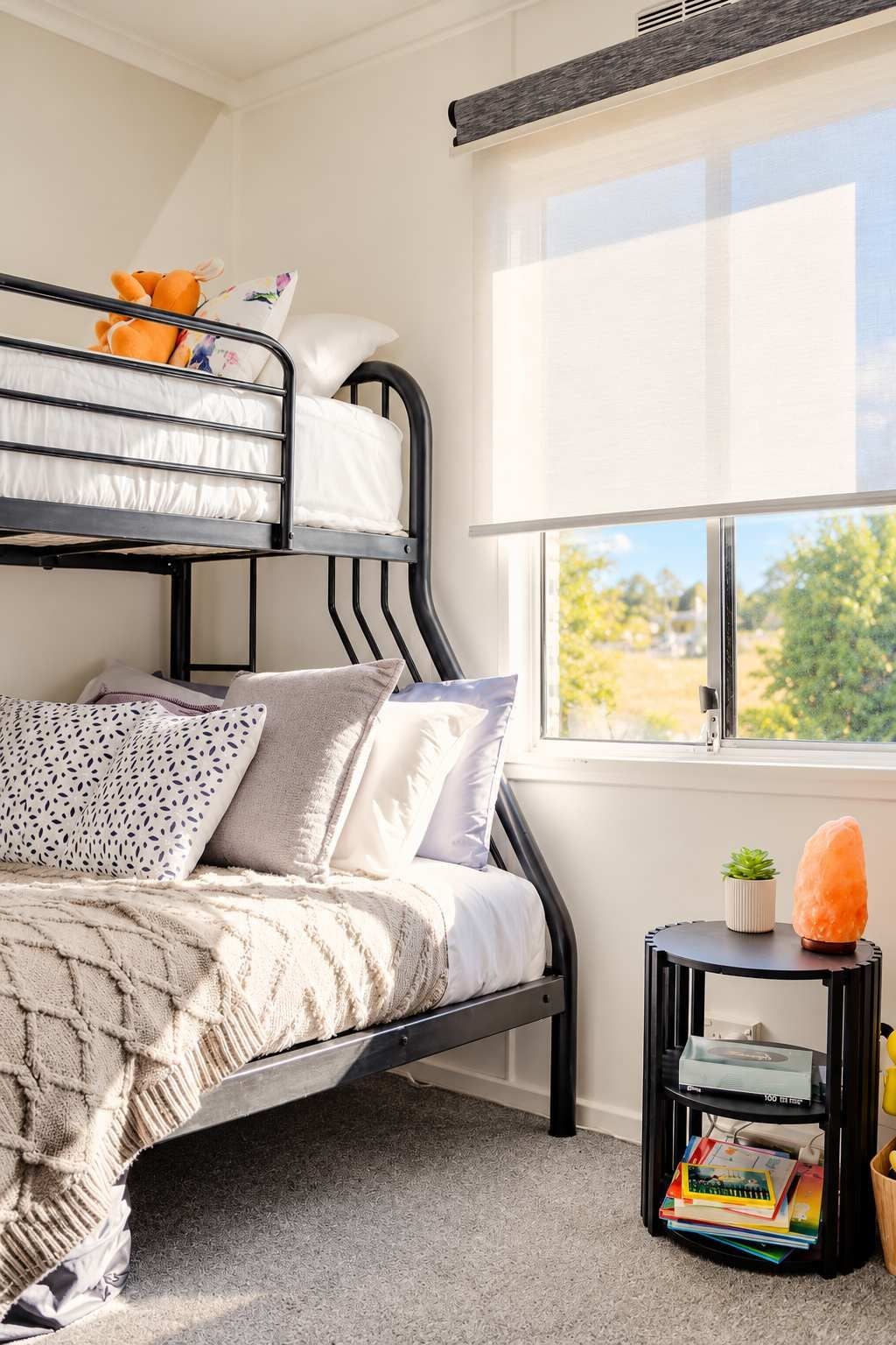 A bunk bed with a window, a small table with a plant and a salt lamp, and a bookshelf.