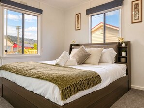 Un dormitorio con una cama grande, dos ventanas, un cabecero y una mesita de noche con un florero.