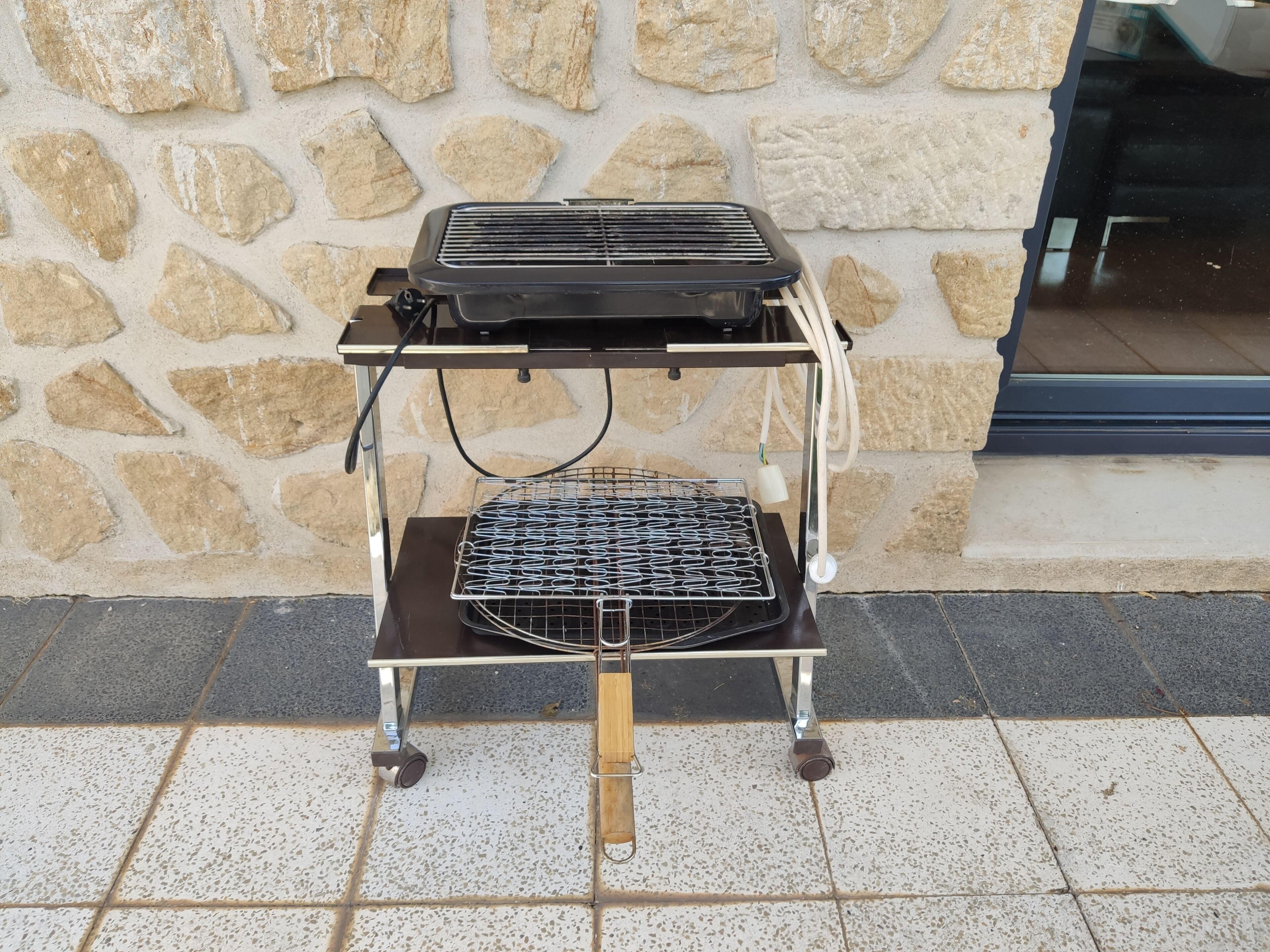 Una parrilla portátil sobre un carrito con ruedas y un muro de piedra de fondo.