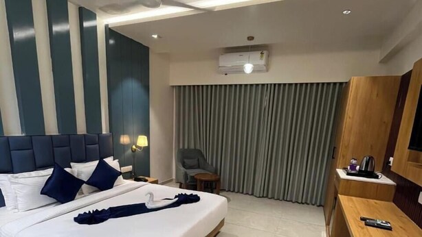 A hotel room with a large bed, a desk, a chair, a ceiling fan, an air conditioning unit, and a window with curtains. - Hotel Premji by Shivalaya Darshanam (Dwarka)