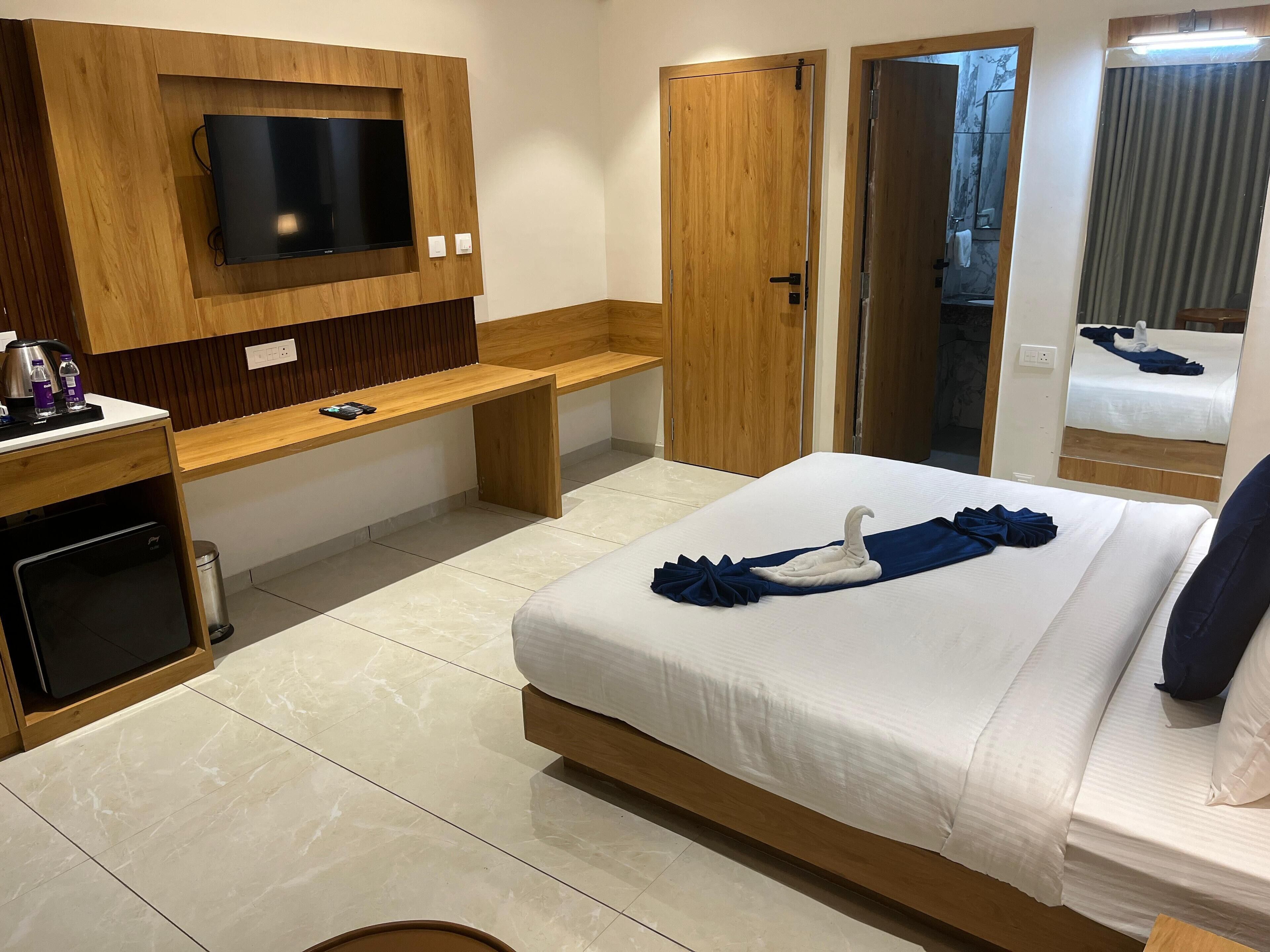 Habitación de hotel con cama, televisor montado en un panel de madera, un banco, escritorio con hervidor y un cubo de basura.