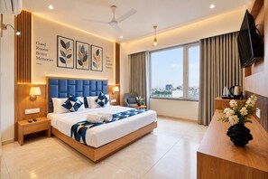 A hotel room with a bed, bedside tables, a desk, a chair, a TV, a window with curtains, and artwork on the walls. - Hotel Premji by Shivalaya Darshanam (Dwarka)