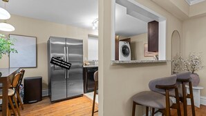 A kitchen with a stainless steel refrigerator, a dishwasher, and a built-in oven. There is a dining area with wooden chairs and a table. - Cozy 1BR Retreat in Central West End with Fireplace, Sofa Bed & City Views (St. Louis)