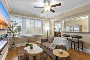 Un salon comprenant un canapé, deux fauteuils, une petite table, un ventilateur de plafond et une grande fenêtre avec des stores.