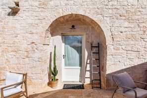 An arched stone doorway leading to a room with a white door, a potted cactus, a metal chair, and a ladder against the wall. - Masseria Maranna Trulli&spa Luxury Villa (Martina Franca)