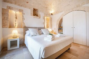A bedroom with a large bed, white linens, a nightstand with a lamp, and a stone wall. - Masseria Maranna Trulli&spa Luxury Villa (Martina Franca)