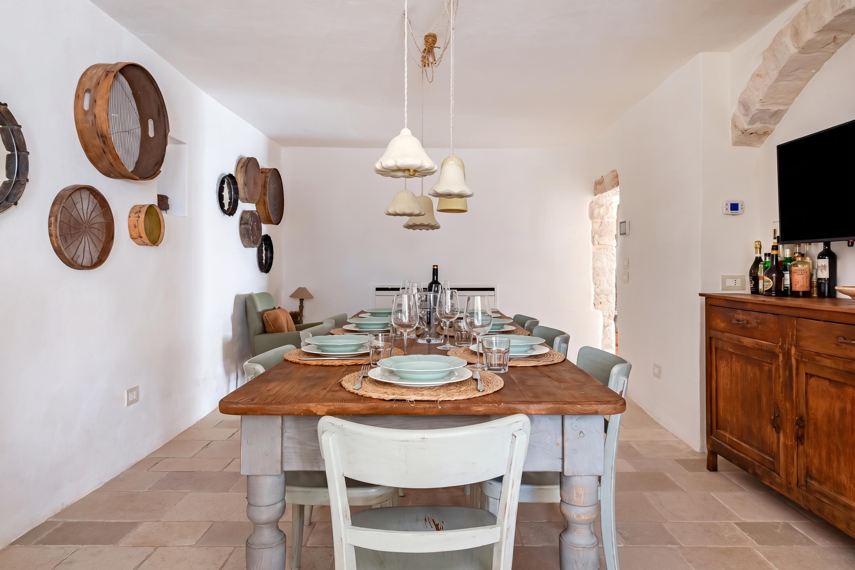A dining area with a wooden table set for four, surrounded by chairs, with a stone wall and a television on the right.