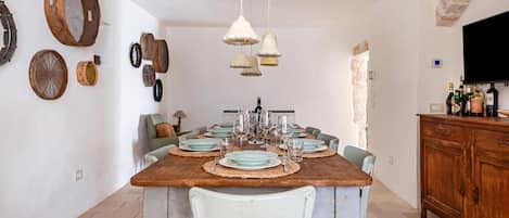 A dining area with a wooden table set for four, surrounded by chairs, with a stone wall and a television on the right.