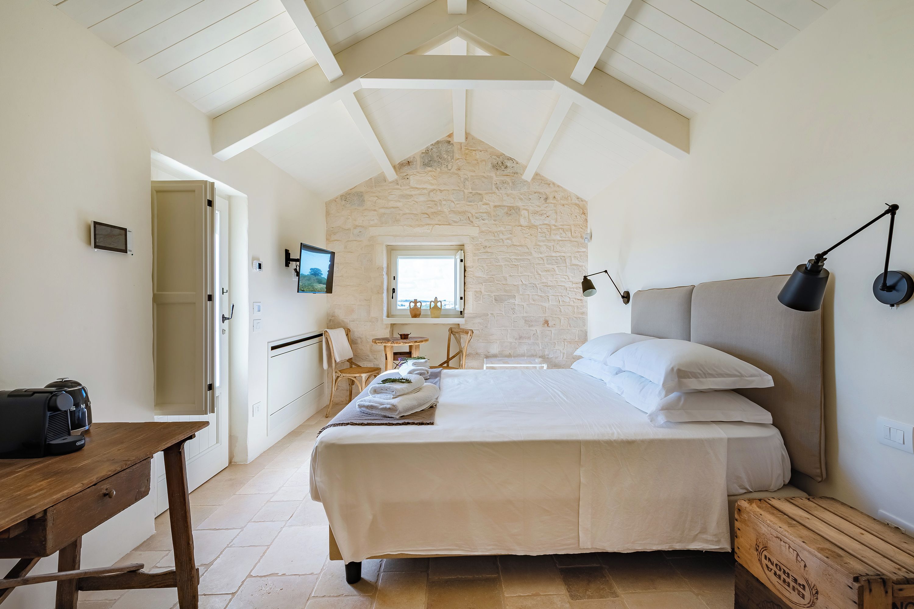 A bedroom with a stone wall, a bed with white linens, a wooden nightstand, a desk with a printer, and a small table with chairs.