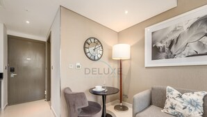 Une chambre d’hôtel moderne avec un canapé gris, un fauteuil gris, une table ronde avec une lampe, une horloge au mur et un tableau encadré.
