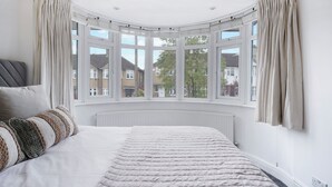 A bedroom with a large bed, a curved bay window, and a view of the neighborhood. - Mount Park Rd Flat (Pinner)