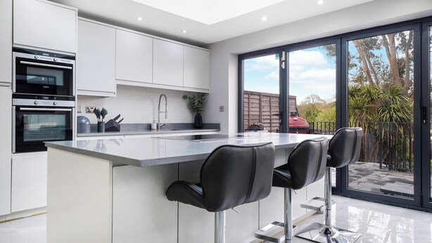 A modern kitchen with a central island, two bar stools, and a large sliding glass door leading to a patio. - Mount Park Rd Flat (Pinner)
