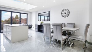 A modern kitchen with a central island, a dining area with a table and chairs, and a large window providing a view of the outdoors. - Mount Park Rd Flat (Pinner)