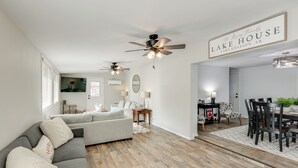 A modern living room with a gray sectional sofa, wooden flooring, ceiling fans, a dining area with a table and chairs, and a sign reading "The Royal Family Lake House". - Bring Your Boat: Family Home Steps to Lake Greeson (Kirby)