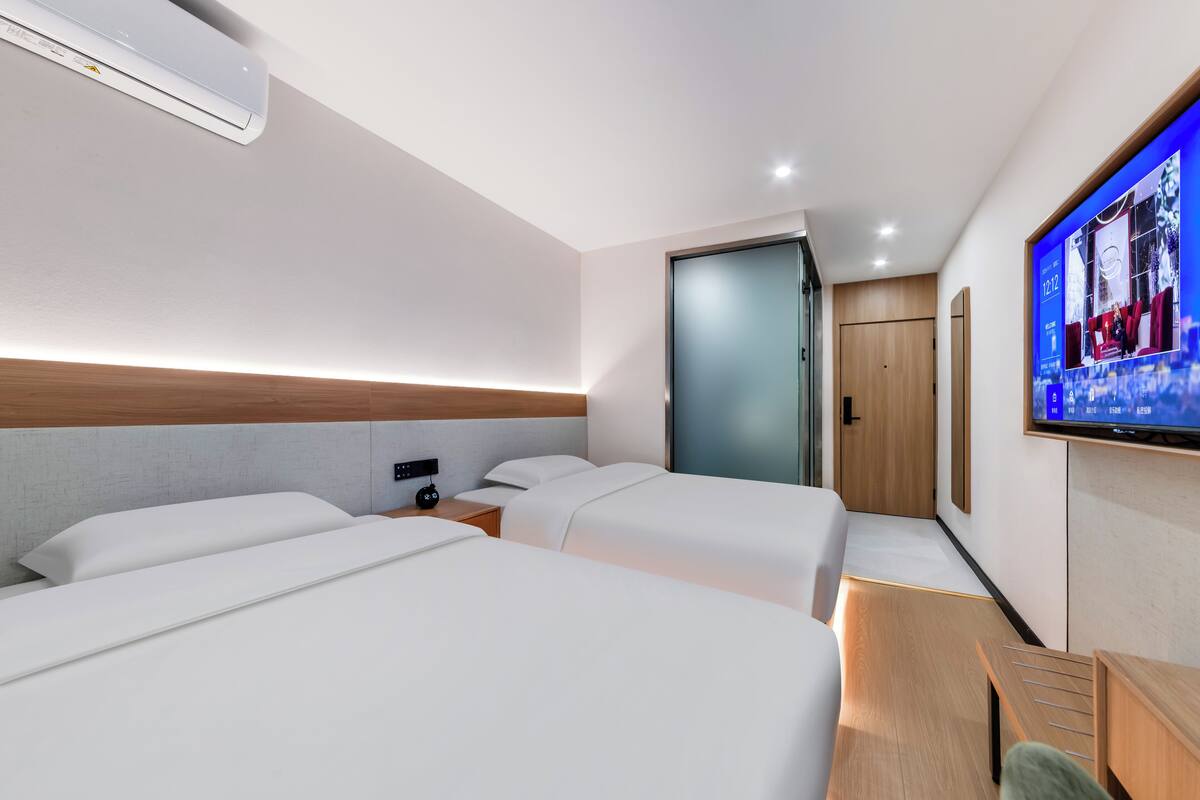 Een moderne hotelkamer met twee bedden, een aan de muur bevestigde televisie, een houten hoofdeinde en een klein bureau met stoel.